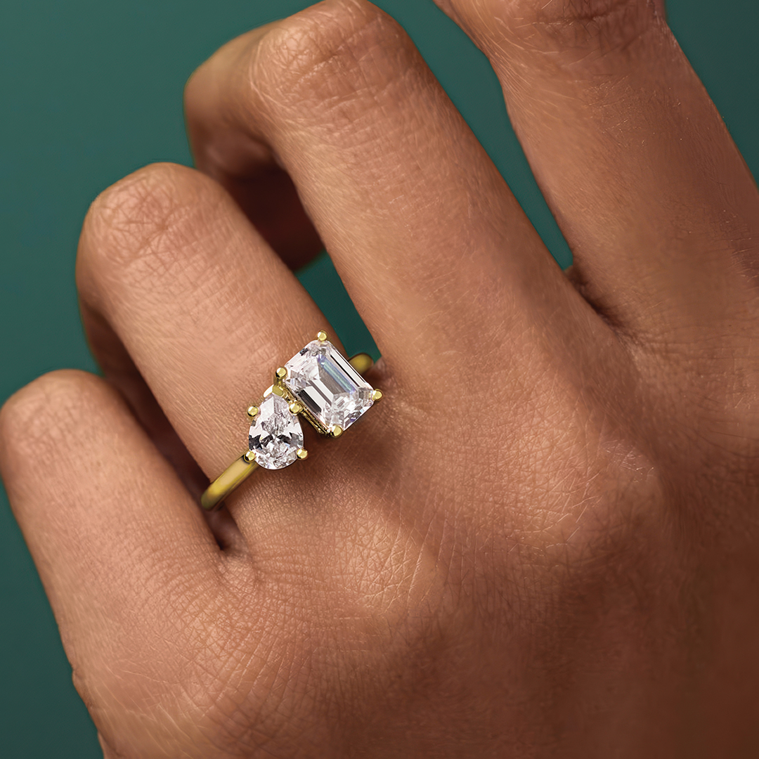 A two stone diamond engagement ring highlights pear and emerald cut stones with a trending yellow gold band. 